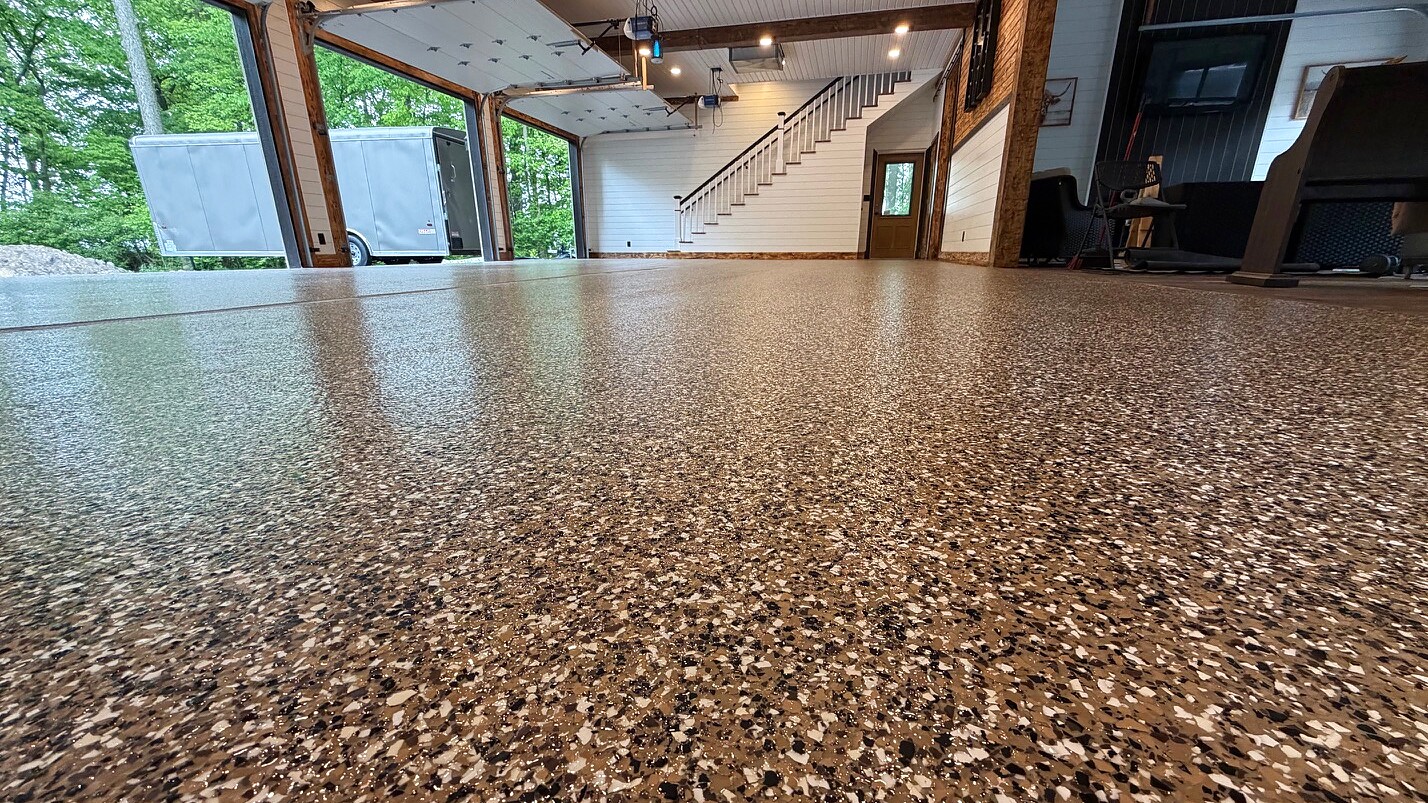 Tan, black, and white flake epoxy floor in a custom residential garage with open bay doors and staircase